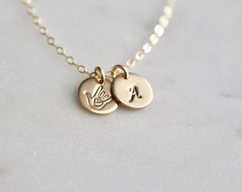 May include: Gold necklace with two charms, one with the American Sign Language symbol for 'I love you' and the other with the letter 'A'.