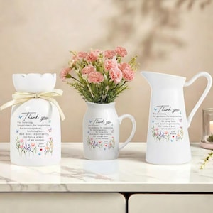 May include: Three white ceramic vases with floral arrangements. The vases have the text "Thank you" and a message. The vase on the left has a ribbon. The middle vase has a handle. The vase on the right is a pitcher.
