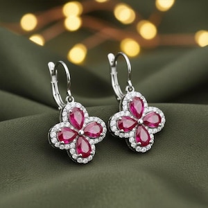 May include: A pair of drop earrings featuring a four-leaf clover design. Each earring contains four pear-cut, deep pink-red stones arranged in a floral pattern, surrounded by a border of small, clear, round-cut stones set in a metallic frame.