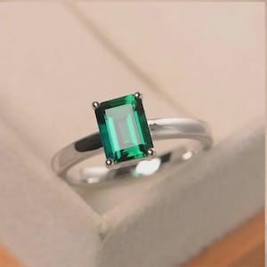 Peut inclure: Une bague argentée ornée d'une pierre précieuse verte taille émeraude fixée par quatre griffes. L'anneau est lisse et poli, présentant une finition métallique nette qui complète la pierre verte rectangulaire vibrante au centre.