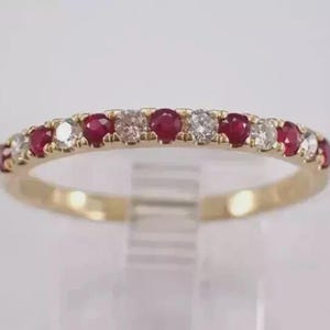 Puede incluir: Un anillo de banda de oro amarillo con un patrón alterno de rubíes rojos y diamantes blancos de corte redondo engastados en una fila a lo largo de la mitad superior. El metal tiene un acabado pulido y las piedras están sujetas con garras.