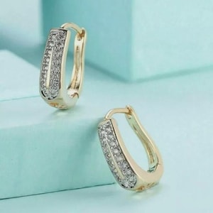 May include: A pair of gold-toned hoop earrings featuring a rectangular, U-shaped design. The front surface of each earring is encrusted with a row of small, clear, sparkling stones that extend along the length of the metal frame.