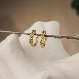 Op de afbeelding: Een paar gouden oorringen, elk versierd met een rij kleine, lichtgroene edelstenen. De oorbellen worden gepresenteerd tegen een neutrale achtergrond, wat hun elegante ontwerp en kleur benadrukt.