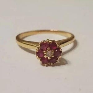 May include: A gold ring featuring a floral-shaped cluster setting. The center of the cluster contains a single small white stone surrounded by six round, deep red rubies. The band is smooth and polished, tapering slightly toward the setting.