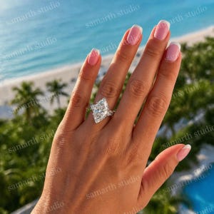 May include: A marquise-cut diamond engagement ring with a halo setting. The ring features a large, elongated diamond as the centerpiece, surrounded by smaller accent stones. The band is also adorned with smaller diamonds. The ring is set against a backdrop of a beach.