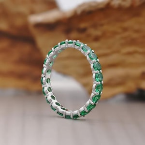 Puede incluir: Un anillo de plata con una banda continua de piedras preciosas verdes de corte ovalado. El engaste del anillo es de color plata brillante, y las piedras están espaciadas uniformemente alrededor de toda la banda. El anillo se muestra sobre un fondo borroso.