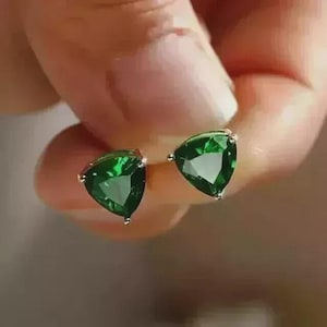May include: A pair of stud earrings featuring faceted, triangle-shaped dark green gemstones set in a metallic frame. The stones have a deep emerald hue with clear, reflective surfaces that catch the light, showcasing a sharp, geometric cut.