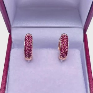 May include: A pair of hoop earrings featuring a rose gold tone metal band encrusted with numerous small, vibrant pink stones. The earrings are positioned upright, showcasing a curved design with a consistent pavé setting across the front surface.