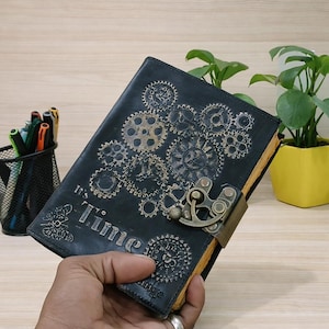May include: Black leather journal with a steampunk gear design and the words "It's Time" embossed on the cover. The journal has a metal clasp and aged paper edges. A unique notebook for writing.