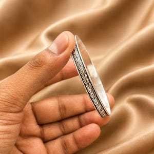 Op de afbeelding: Een zilveren armband met een gedetailleerd bladerenpatroon. De armband wordt tegen een goudkleurige stoffen achtergrond gehouden, wat het ingewikkelde ontwerp en de gepolijste afwerking van het sieraad laat zien.