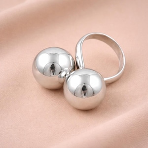 以下が含まれることがあります： 光沢のある銀色の金属製の球体が2つ、リングに繋がっています。球体は非常に反射性が高く、表面は滑らかです。リングも銀色で、曲線的なデザインです。背景は柔らかいピンク色の布です。