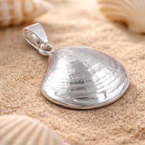 Könnte beinhalten: Silberner Muschel-Anhänger mit strukturierter Oberfläche, der auf Sand liegt. Der Anhänger ist eine detaillierte Nachbildung einer Muschelschale mit einer Schlaufe für eine Kette. Weitere Muscheln befinden sich im Hintergrund.