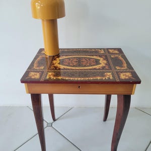 Peut inclure: Petite table d'appoint ancienne avec un dessus en bois incrusté et un seul tiroir. La table a quatre pieds incurvés et une lampe jaune moutarde. La lampe a un abat-jour arrondi et une base cylindrique. La table mesure environ 60 cm de haut.