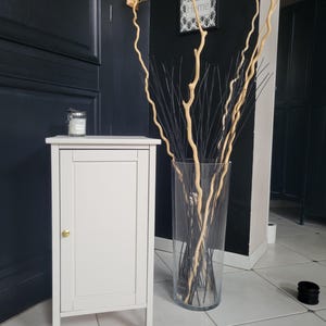 May include: A light gray rectangular cabinet with a single door and a small gold knob, topped with a glass candle jar. Beside it stands a tall, clear glass vase filled with long, thin, light-colored curly branches and dark, straight decorative sticks.