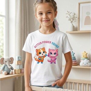 Pode incluir: Camiseta branca com o texto "Kitten Power Squad" e dois gatinhos de desenho animado. Um gatinho &eacute; laranja e o outro rosa. O gatinho laranja est&aacute; usando uma roupa laranja e azul, e o gatinho rosa est&aacute; usando uma roupa rosa.