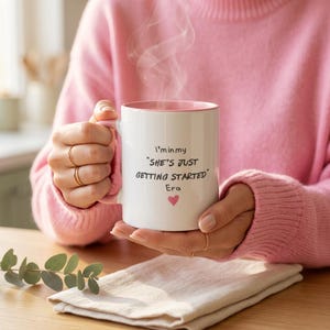 Può includere: Una tazza di ceramica bianca con interno rosa, tenuta da una persona con un maglione rosa. La tazza ha la scritta "I'm in my 'SHE'S JUST GETTING STARTED' Era" con un cuore rosa. Il vapore sale dalla tazza.