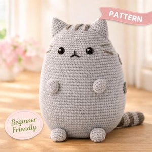 May include: A grey crocheted cat plush toy with dark grey stripes on its head and tail, black plastic eyes, and a small embroidered nose and mouth. A pink banner at the top reads PATTERN, and a pink oval label at the bottom reads Beginner Friendly.