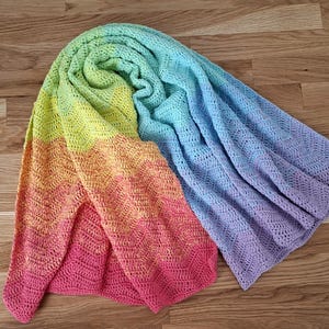 May include: A crochet blanket featuring a vibrant rainbow gradient pattern. The textile displays a chevron stitch design transitioning through shades of pink, orange, yellow, light green, light blue, and purple in a soft, textured yarn construction.