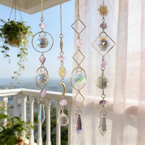 Puede incluir: Tres atrapasoles colgantes con cadenas doradas, formas geométricas y cristales. Cada uno presenta bolas de cristal transparente, piedras de amatista y detalles metálicos dorados, incluyendo amuletos de sol y luna. El fondo muestra un balcón y vegetación.