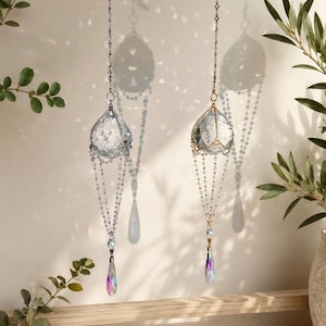 Puede incluir: Dos atrapasoles de cristal colgando de cadenas. Uno tiene una cadena plateada y el otro una cadena dorada. Cada uno tiene un colgante de cristal facetado en forma de l&aacute;grima con colores iridiscentes. Cuelgan frente a una pared blanca.
