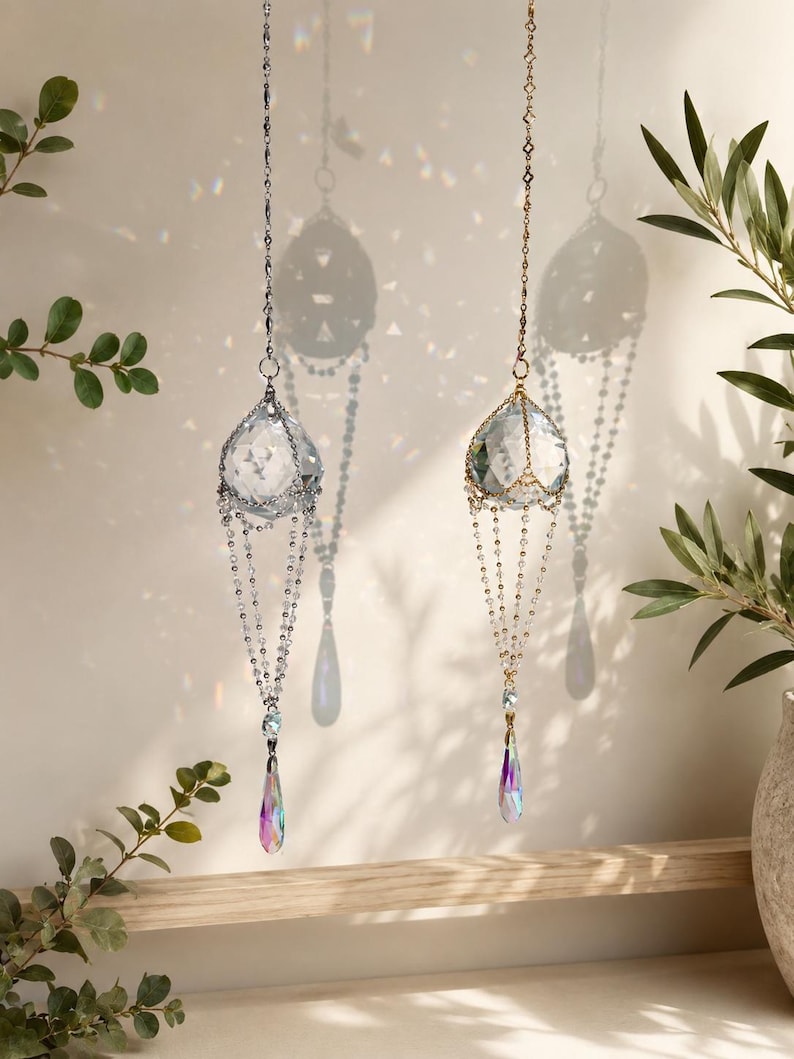 Puede incluir: Dos atrapasoles de cristal colgando de cadenas. Uno tiene una cadena plateada y el otro una cadena dorada. Cada uno tiene un colgante de cristal facetado en forma de l&aacute;grima con colores iridiscentes. Cuelgan frente a una pared blanca.