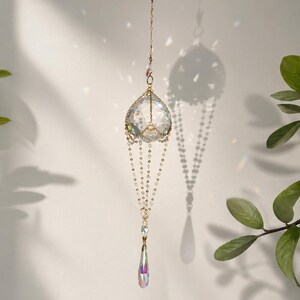 Puede incluir: Un atrapasoles de cristal colgante con un marco y cadena de metal dorado. El atrapasoles presenta una gran esfera de cristal facetado en la parte superior, con cuentas de cristal m&aacute;s peque&ntilde;as y un colgante de cristal en forma de l&aacute;grima debajo.