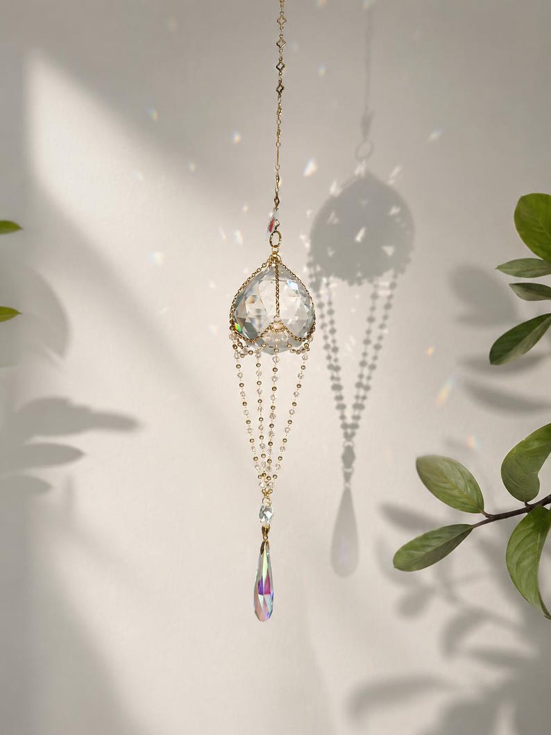 Puede incluir: Un atrapasoles de cristal colgante con un marco y cadena de metal dorado. El atrapasoles presenta una gran esfera de cristal facetado en la parte superior, con cuentas de cristal m&aacute;s peque&ntilde;as y un colgante de cristal en forma de l&aacute;grima debajo.