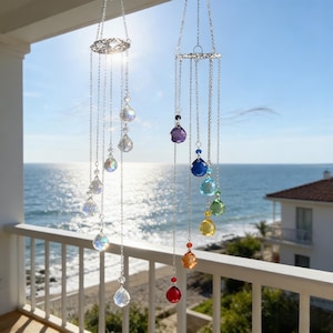 May include: Two crystal wind chimes hanging on a balcony overlooking the sea. Each chime has a decorative silver top with multiple strands of clear and colourful crystal beads. The sun shines brightly in the background, reflecting off the water.