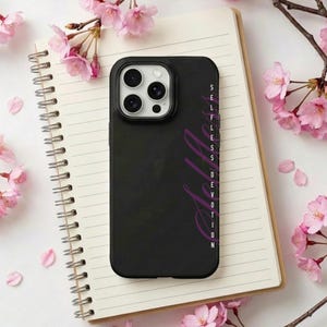 Op de afbeelding: Zwarte telefoonhoes met de woorden "Selfless Devotion" in paars. De hoes ligt op een wit notitieboek, omringd door roze kersenbloesemtakken en bloemblaadjes.