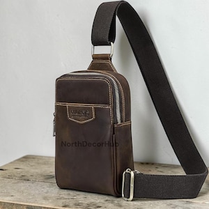 May include: A brown leather sling bag with a long, adjustable strap. The bag has a vertical design with a zippered main compartment and a smaller front pocket. The text "CURUSO" is visible on the front. The bag is displayed on a wooden surface.