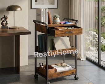May include: A rolling storage cart with a dark metal frame and rustic brown wood-grain shelves. It features a top drawer, two open lower shelves, and four caster wheels. The cart holds books, a small potted succulent, and office supplies.