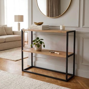 May include: A two-tier console table with a light wood top and a black metal frame. The top shelf holds books, a small bowl, and a candle. A potted plant and a magnifying glass are on the lower shelf. The table is in a living room setting.