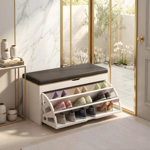 May include: A white shoe storage bench with a dark grey cushioned seat. The bench has three angled shelves filled with various shoes. The bench is in a well-lit entryway with a marble wall and a view of the outdoors.