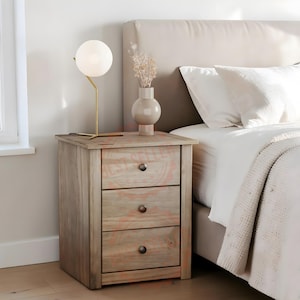 May include: A light brown wooden bedside table with three drawers, each featuring a single dark metal knob. A spherical white glass lamp with a thin gold-toned stand and a beige ceramic vase containing dried flowers sit on the top surface.