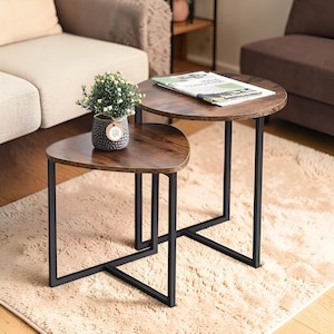 May include: Two nesting tables with dark brown wooden tops and black metal frames. One table is round, the other is a rounded triangle shape. A small potted plant sits on the smaller table. The tables are on a beige rug.
