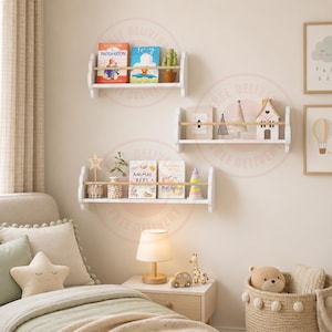 May include: Three white wall-mounted floating shelves with light wood rails, displaying children's books, a small potted cactus, a wooden house ornament, a star-shaped sign, and a colourful stacking toy, arranged on a light-coloured bedroom wall.