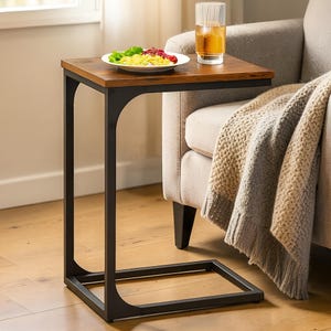 May include: A brown and black side table with a C-shaped design. The rectangular wooden tabletop holds a plate of food and a glass of iced tea. A beige blanket is draped over a light-coloured sofa. The table is approximately 60 cm high.