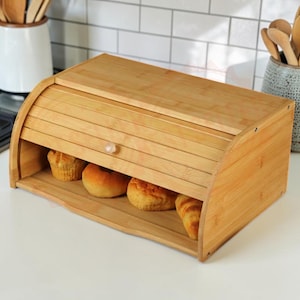 May include: A rectangular bamboo bread bin with a curved, sliding roll-top door and a small round knob. The interior of the wooden container holds four baked goods, including a muffin, a round roll, and two other pieces of golden-brown bread.