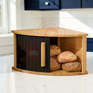 May include: A corner bread bin with a light wood frame and a black sliding tambour door featuring a vertical wooden handle. The interior holds three assorted artisan bread loaves with visible seeds and crusty textures on a wooden base.