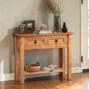 May include: A light brown wooden console table featuring two drawers with black ring pulls, a flat rectangular top, and a lower shelf. The table holds two small picture frames, a stack of books, a glass vase with dried plants, and a dark vase.