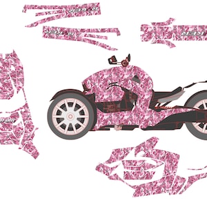 May include: Pink camouflage-patterned Can-Am Ryker three-wheeled motorcycle with matching decal set. The Ryker has black accents, white wheels, and the word "Ryker" in white. Additional decals are shown separately.