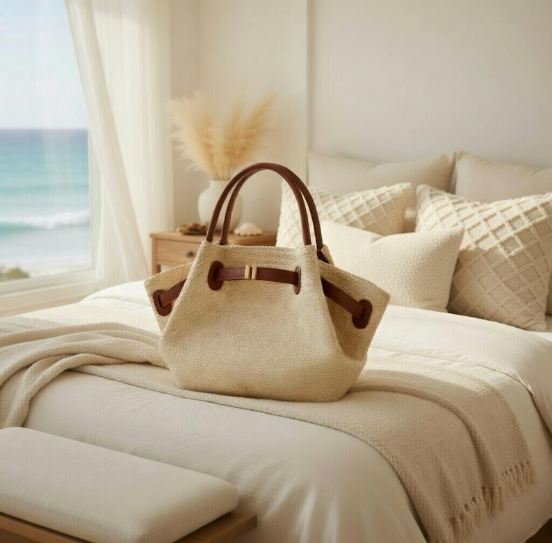 Puede incluir: Un bolso tote tejido beige con asas y detalles de cuero marr&oacute;n descansa sobre una cama. El bolso tiene forma rectangular, parte superior fruncida y una hebilla decorativa. El fondo muestra una ventana con vistas al mar.