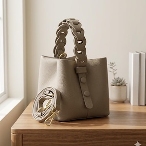 Può includere: Una borsa a secchiello color tortora con superficie testurizzata, caratterizzata da un manico unico a maglie circolari e un dettaglio a cinturino verticale con due bottoni abbinati. La borsa include una tracolla regolabile e rimovibile con chiusure dorate.