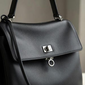 Pu&ograve; includere: Una borsa in pelle nera con chiusura a patta e chiusura argentata. La borsa ha un manico superiore e una tracolla staccabile. La pelle sembra liscia e la borsa ha una forma strutturata.