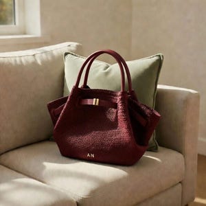 Puede incluir: Un bolso tote burdeos con una textura tejida, con una asa superior y un cierre de hebilla dorada. El bolso tiene las iniciales "AN" en dorado en la parte delantera. El bolso est&aacute; sobre un sof&aacute; beige con un coj&iacute;n verde.