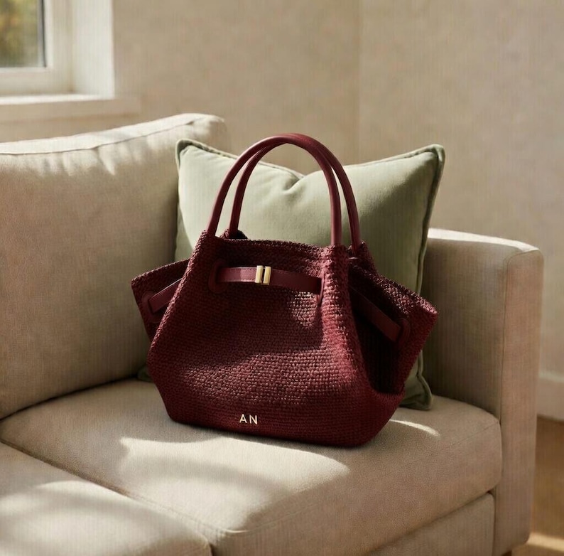 Puede incluir: Un bolso tote burdeos con una textura tejida, con una asa superior y un cierre de hebilla dorada. El bolso tiene las iniciales "AN" en dorado en la parte delantera. El bolso est&aacute; sobre un sof&aacute; beige con un coj&iacute;n verde.