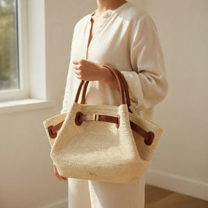 Puede incluir: Un bolso tote tejido beige con asas y detalles de cuero marr&oacute;n. El bolso tiene un cierre de cord&oacute;n y una forma rectangular. El bolso lo sostiene una persona que lleva una camisa color crema.