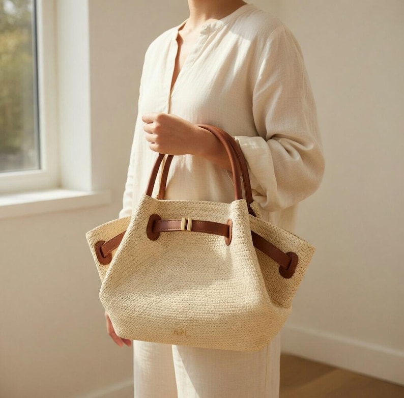 Puede incluir: Un bolso tote tejido beige con asas y detalles de cuero marr&oacute;n. El bolso tiene un cierre de cord&oacute;n y una forma rectangular. El bolso lo sostiene una persona que lleva una camisa color crema.