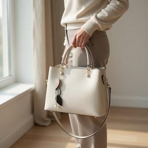 May include: Cream-colored handbag with a structured design, featuring top handles and a detachable shoulder strap. The bag is adorned with gold-toned hardware and decorative leaf charms. A pearl accent hangs from the handle. The bag is held by a person wearing neutral-toned clothing.