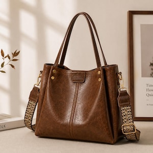 Peut inclure: Un sac cabas marron foncé à la finition texturée, doté de deux anses supérieures et d'une bandoulière tissée à motifs amovible. Le sac est agrémenté de finitions en métal doré, incluant des rivets à la base des anses et des clips latéraux.